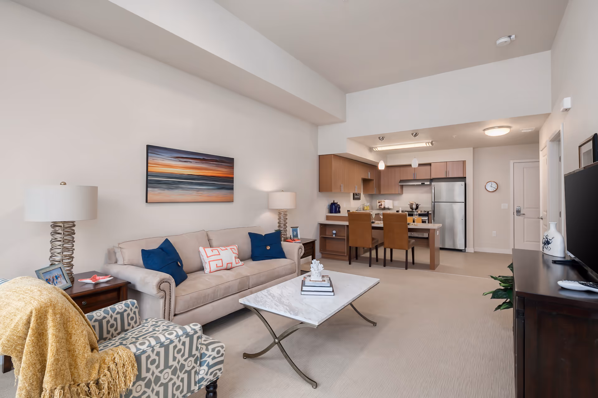 A modern living room and kitchen area in an apartment. The living room features a beige sofa with blue and patterned pillows, a patterned armchair with a yellow throw blanket, two table lamps, a coffee table with books and decor, and a wall-mounted painting of a sunset over the ocean. The kitchen has wooden cabinets, a stainless steel refrigerator, a stove, and a kitchen island with two brown chairs. The walls are light-colored and the floor is carpeted.