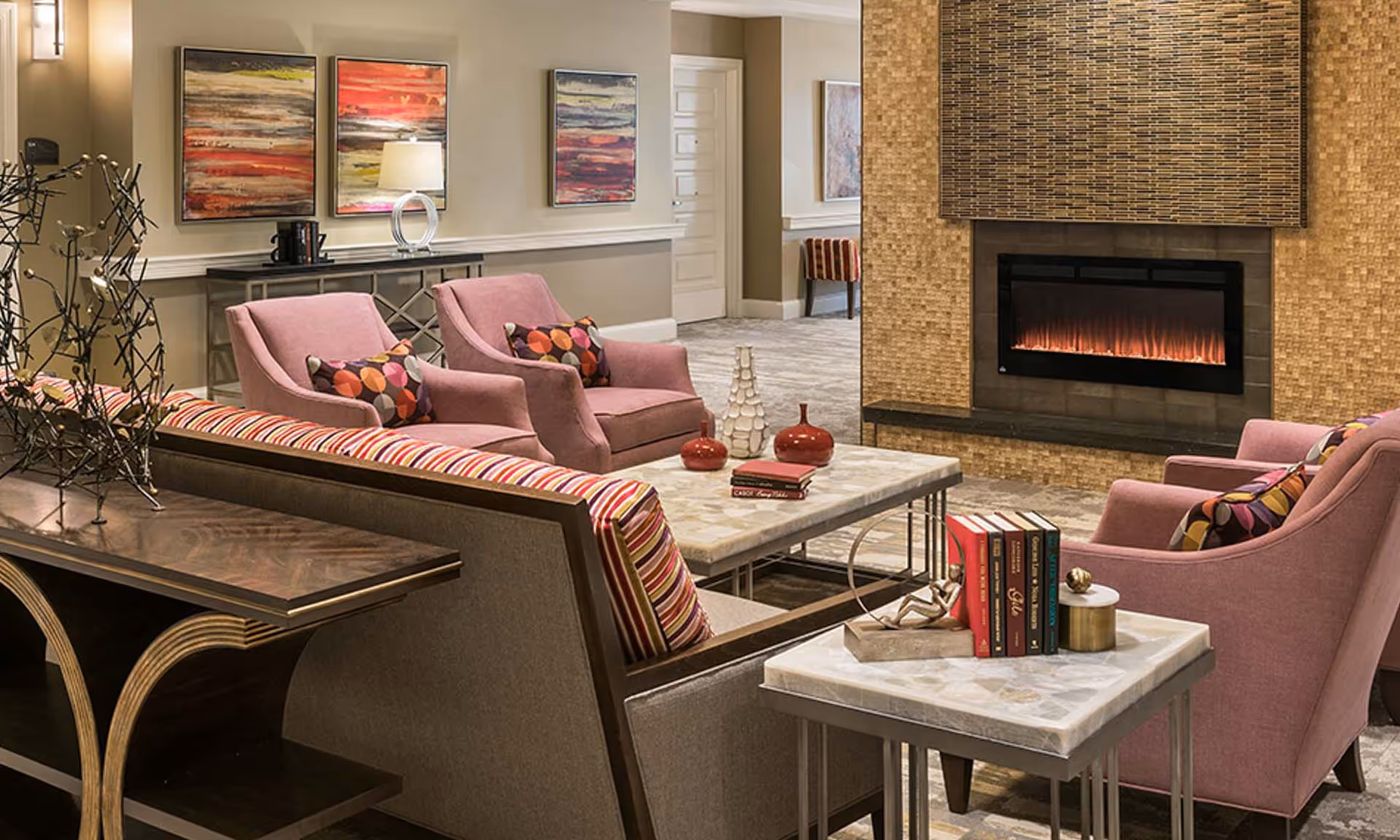A cozy living room area with a modern electric fireplace set in a textured tile wall. The room features two pink upholstered armchairs with colorful patterned pillows, a sofa with striped cushions, and two marble-top tables. Decorative items including books, vases, and a small sculpture are placed on the tables. The walls are adorned with abstract paintings and a console table with a lamp and coffee maker is visible in the background.
