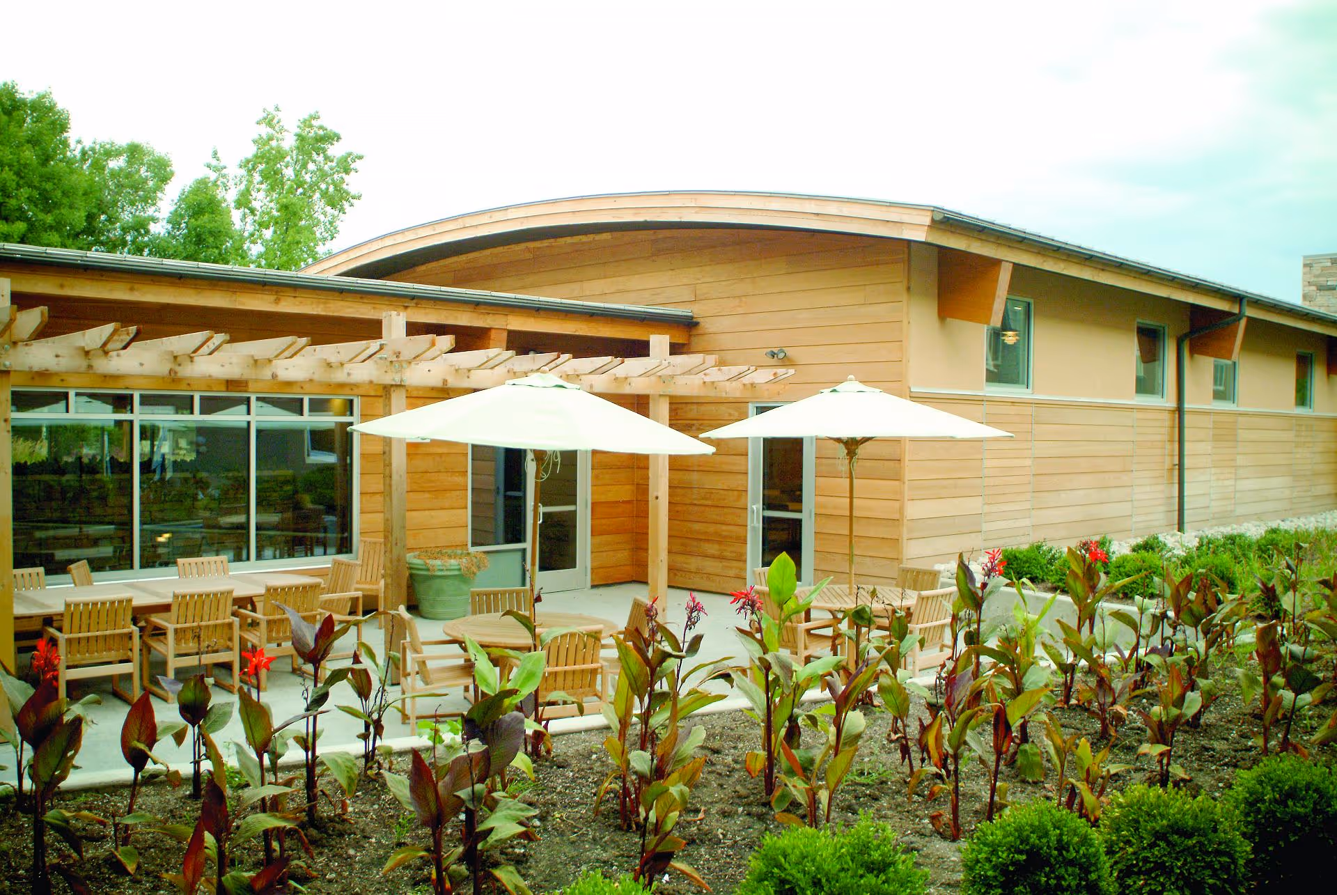 Outdoor patio area at John Knox Village featuring wooden chairs and tables with large white umbrellas, surrounded by a garden with green and red plants. The building has wooden siding and large windows.