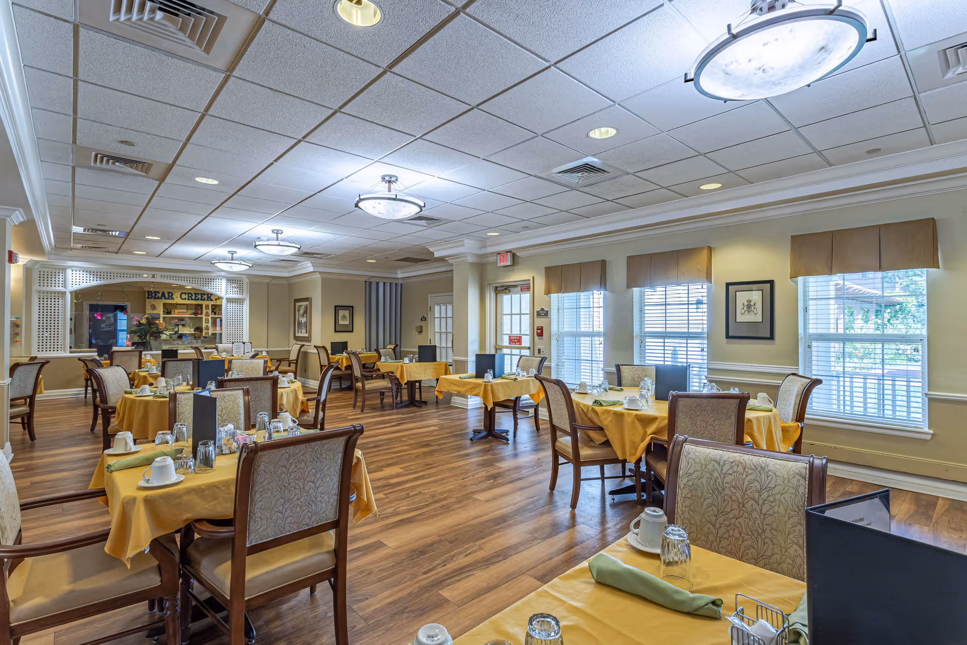 A bright and spacious dining room in Bear Creek Assisted & Senior Living with multiple tables covered in yellow tablecloths, each set with cups, glasses, napkins, and menus. The room has wooden flooring, large windows with blinds, and ceiling lights. A service window with the Bear Creek sign is visible in the background.