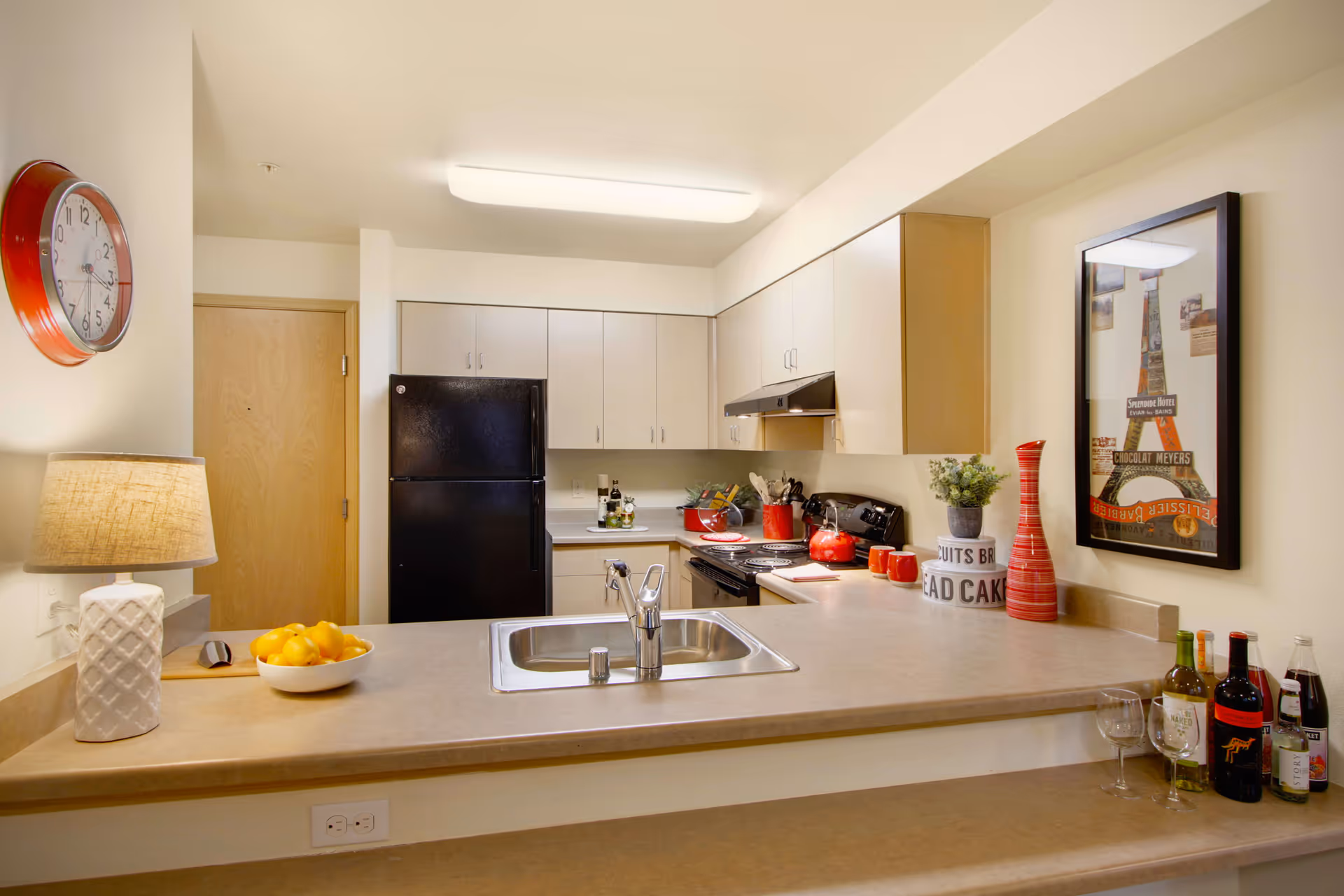 A modern kitchen with beige countertops and light wood cabinets. The kitchen features a black refrigerator, a black stove with a range hood, a stainless steel sink, and various kitchen items including a bowl of lemons, a lamp, red kitchen utensils, a red vase, and bottles of wine and soda. A wall clock and a framed picture are also visible on the walls.
