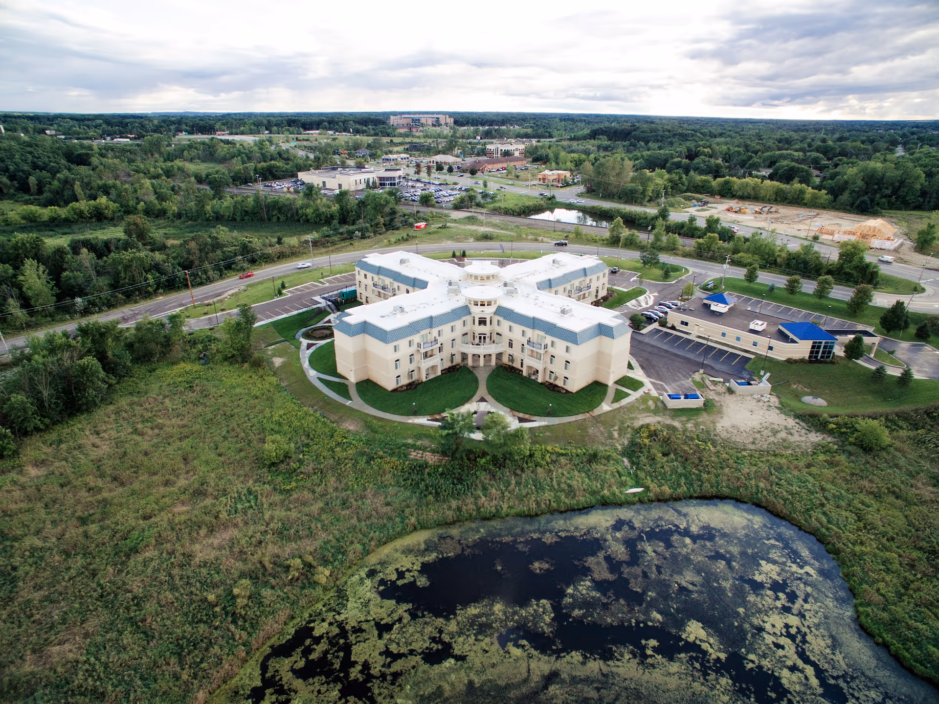 Aerial view of a cross-shaped senior living facility surrounded by parking areas, roads, green fields, and a nearby pond.