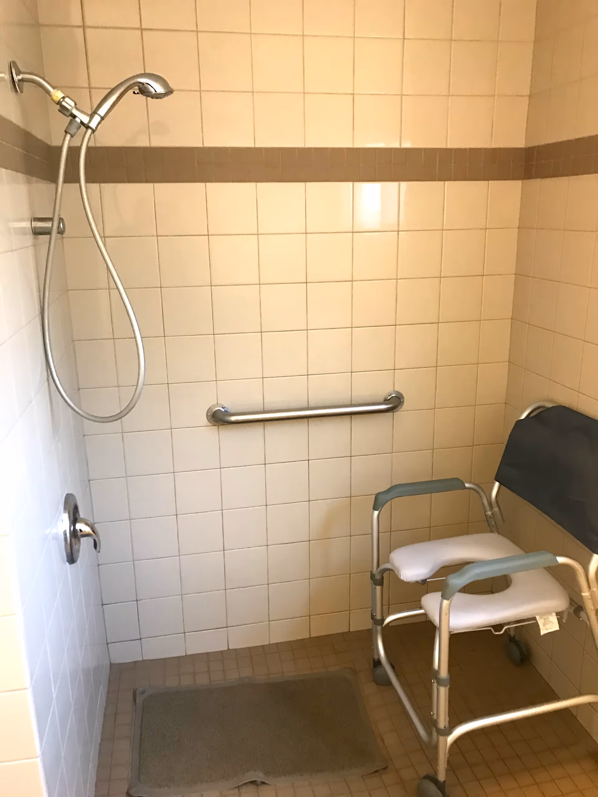 A tiled shower area with beige and brown tiles, featuring a handheld showerhead attached to the wall, a horizontal grab bar, a gray bath mat on the floor, and a shower chair with armrests and wheels.