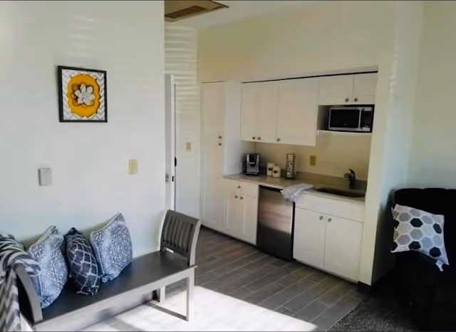 A small kitchenette area with white cabinets, a microwave, a coffee maker, and a sink. To the left, there is a bench with several patterned cushions and a chair. A framed yellow and white flower artwork hangs on the wall above the bench. The floor is tiled and sunlight is streaming into the room.