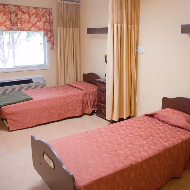A room with two single beds covered in matching reddish bedspreads. There is a window with a red and white patterned valance, a small wooden nightstand between the beds, and beige privacy curtains hanging from the ceiling. The walls are light-colored and the floor is carpeted.