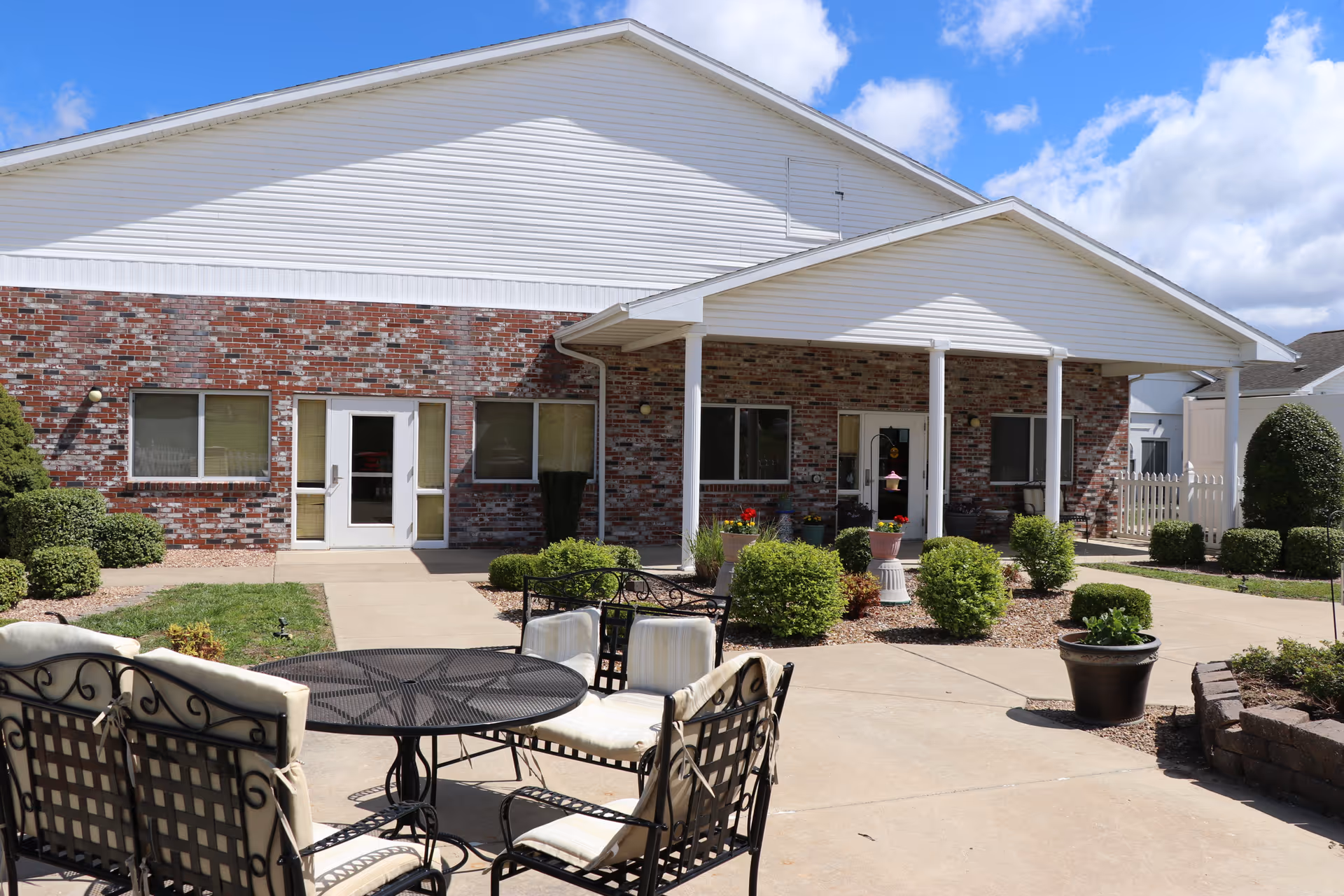 Patio and landscaped courtyard in front of a brick and white-sided building with a covered entrance and outdoor seating.