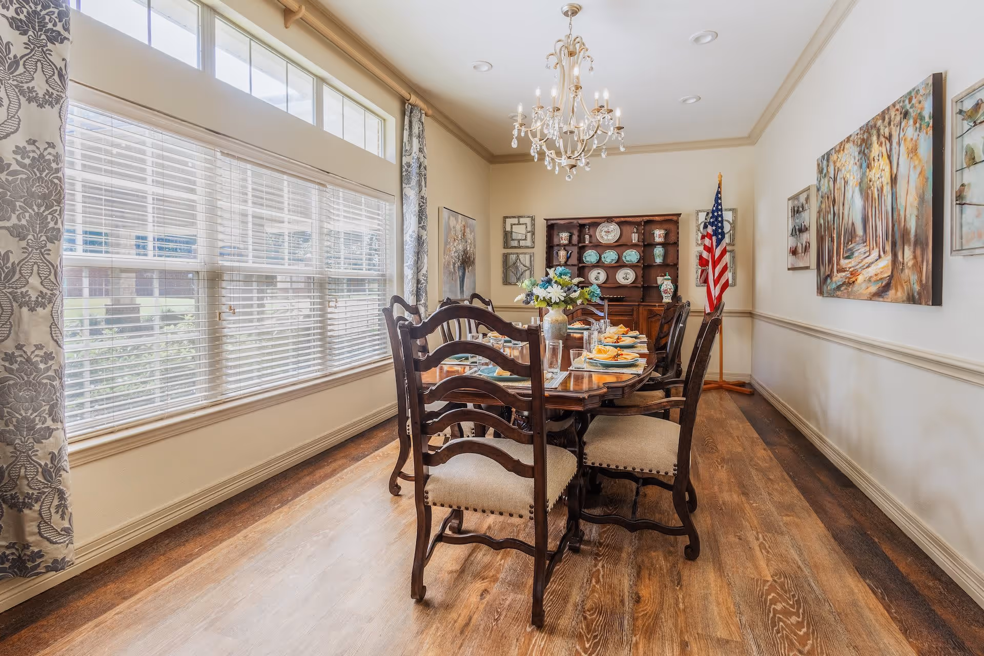 A bright dining room with a wooden dining table set for a meal, surrounded by six cushioned wooden chairs. The room features large windows with white blinds and patterned curtains, a chandelier hanging from the ceiling, a wooden cabinet displaying decorative plates and vases, an American flag in the corner, and artwork on the walls.