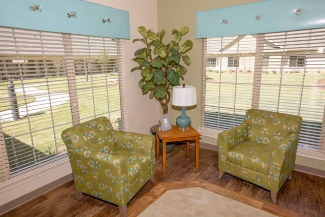 A cozy sitting area with two green armchairs featuring a floral pattern, positioned around a small wooden side table with a blue lamp and a framed photo. Behind the chairs are large windows with blinds and blue valances, and a tall leafy plant is placed in the corner. The floor is wooden with a beige rug partially visible.
