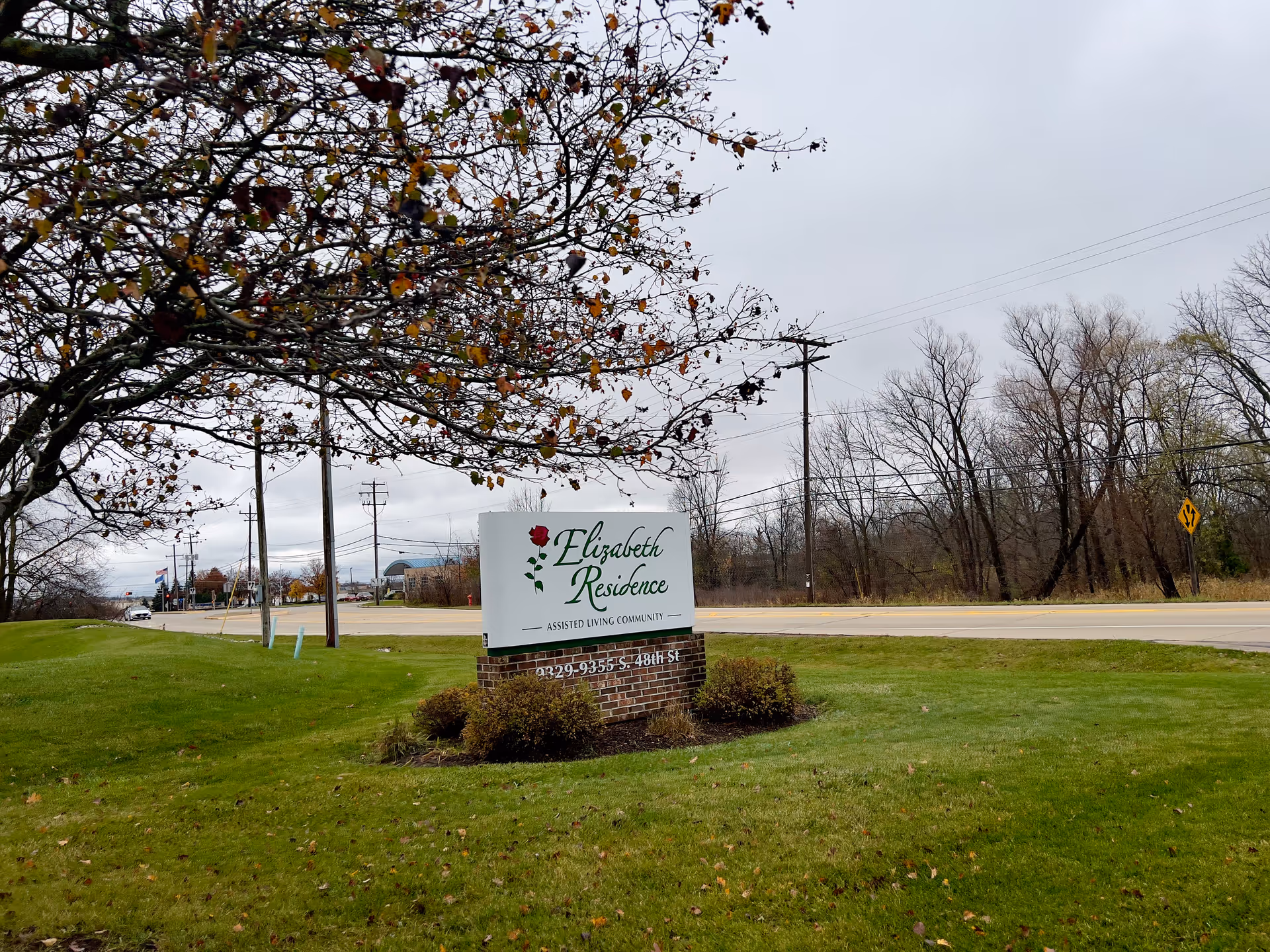 A roadside sign for an assisted living community stands on a grassy lawn with trees and a road in the background.