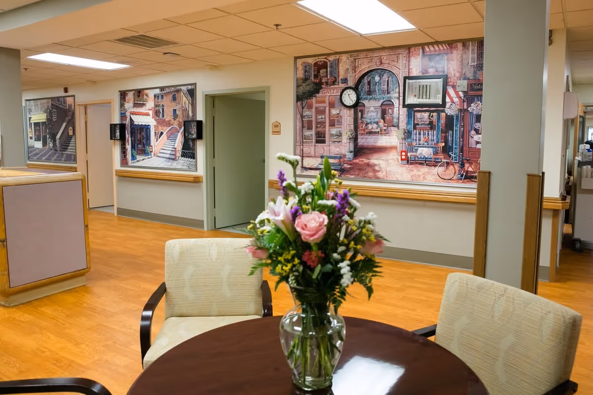 Interior view of a senior living facility hallway with wooden flooring and light-colored walls. There is a round table with a vase of colorful flowers in the foreground and two upholstered chairs. The walls are decorated with large paintings depicting street scenes, and there is a clock mounted on the wall. Several doorways and a reception desk area are visible in the background.