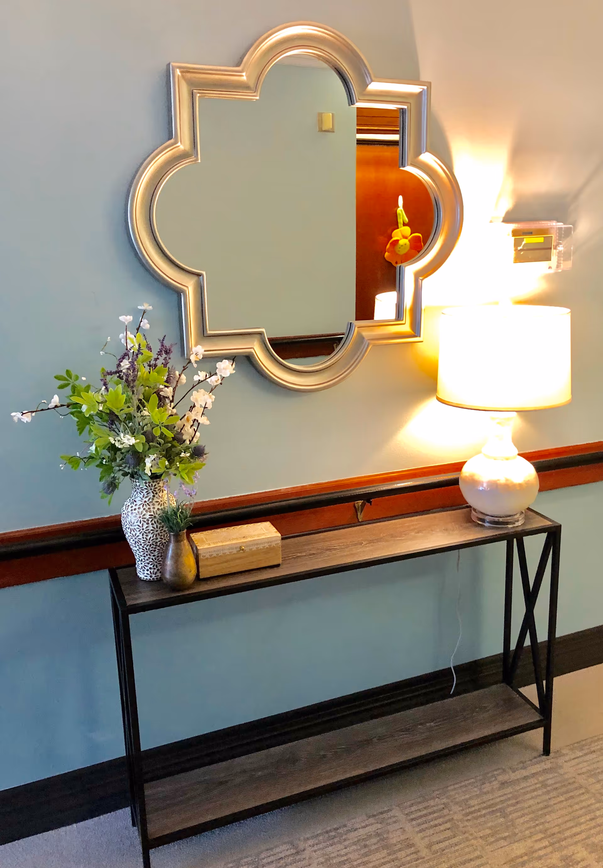 A hallway console table with a decorative quatrefoil mirror, vase of flowers, and a lit table lamp against a light blue wall.