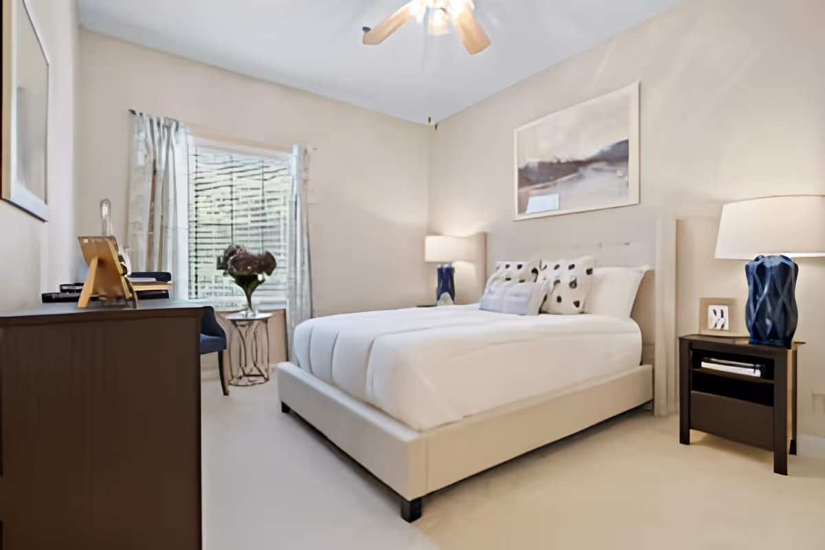 A bright bedroom with a large bed featuring white bedding and decorative pillows. There are two bedside tables with blue lamps, a dresser with framed photos and decor, a chair, and a window with sheer curtains allowing natural light to enter. A ceiling fan is visible above.