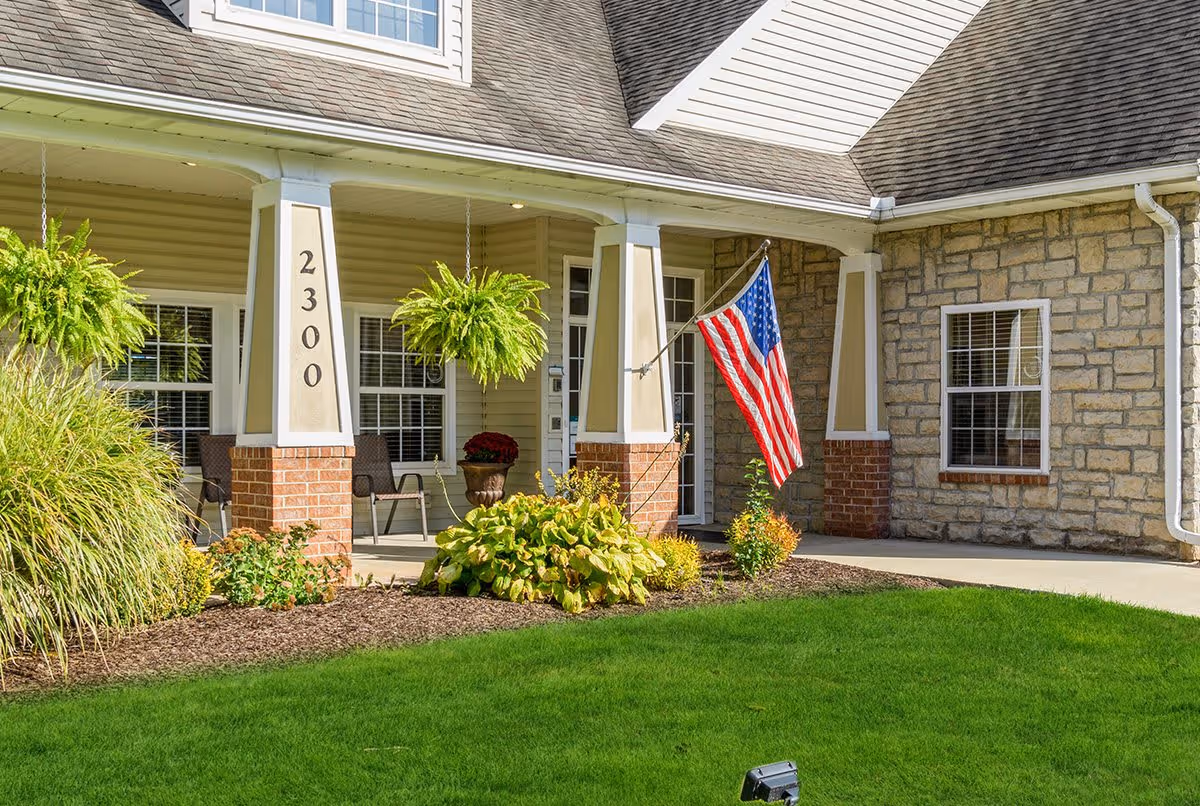 Front porch of a building with beige siding and stone walls, featuring two white columns with brick bases, hanging green plants, an American flag mounted on one column, and a well-maintained green lawn with landscaping in front.