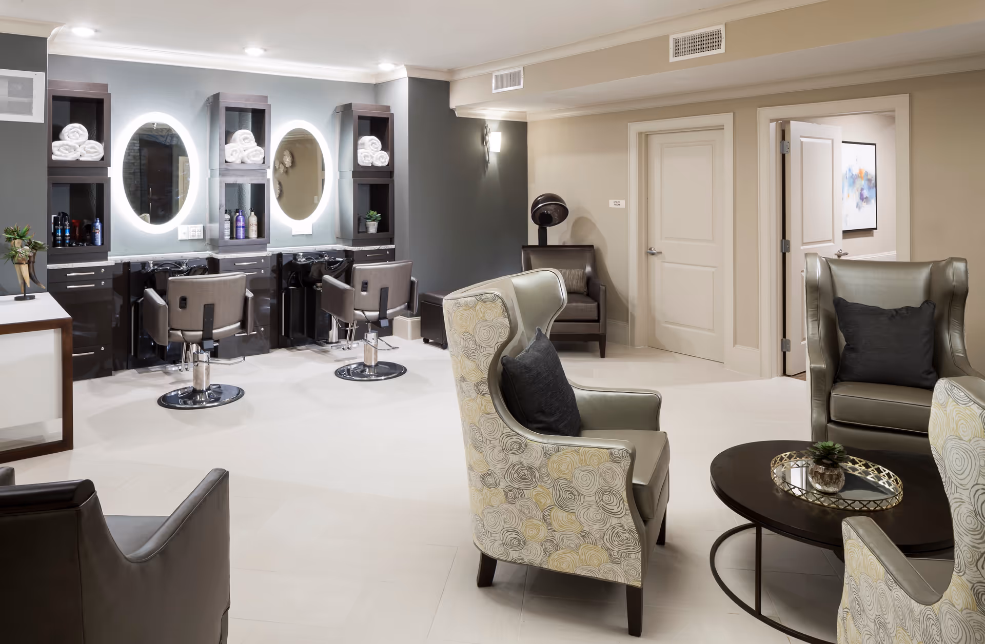 A modern salon area in a senior living facility featuring two styling stations with illuminated oval mirrors, salon chairs, and shelves stocked with towels and hair products. In the foreground, there is a seating area with patterned armchairs, a round coffee table with a small plant, and additional seating in the background near closed doors.