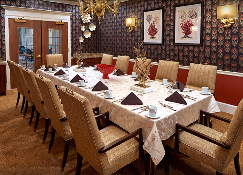 A formal dining room with a long rectangular table covered with a white patterned tablecloth. The table is set with white cups, saucers, silverware, and dark folded napkins. There are twelve upholstered chairs around the table. The walls have a dark patterned wallpaper with framed coral artwork and gold wall sconces. Double wooden doors with glass panels are visible in the background.