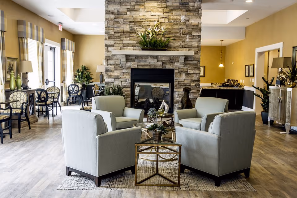 A cozy living room area in an assisted living facility featuring four light gray armchairs arranged around a glass coffee table with decorative plants. The room has a stone fireplace with a plant on the mantel, wood flooring, and large windows with striped curtains letting in natural light. Additional seating and lamps are visible along the walls.