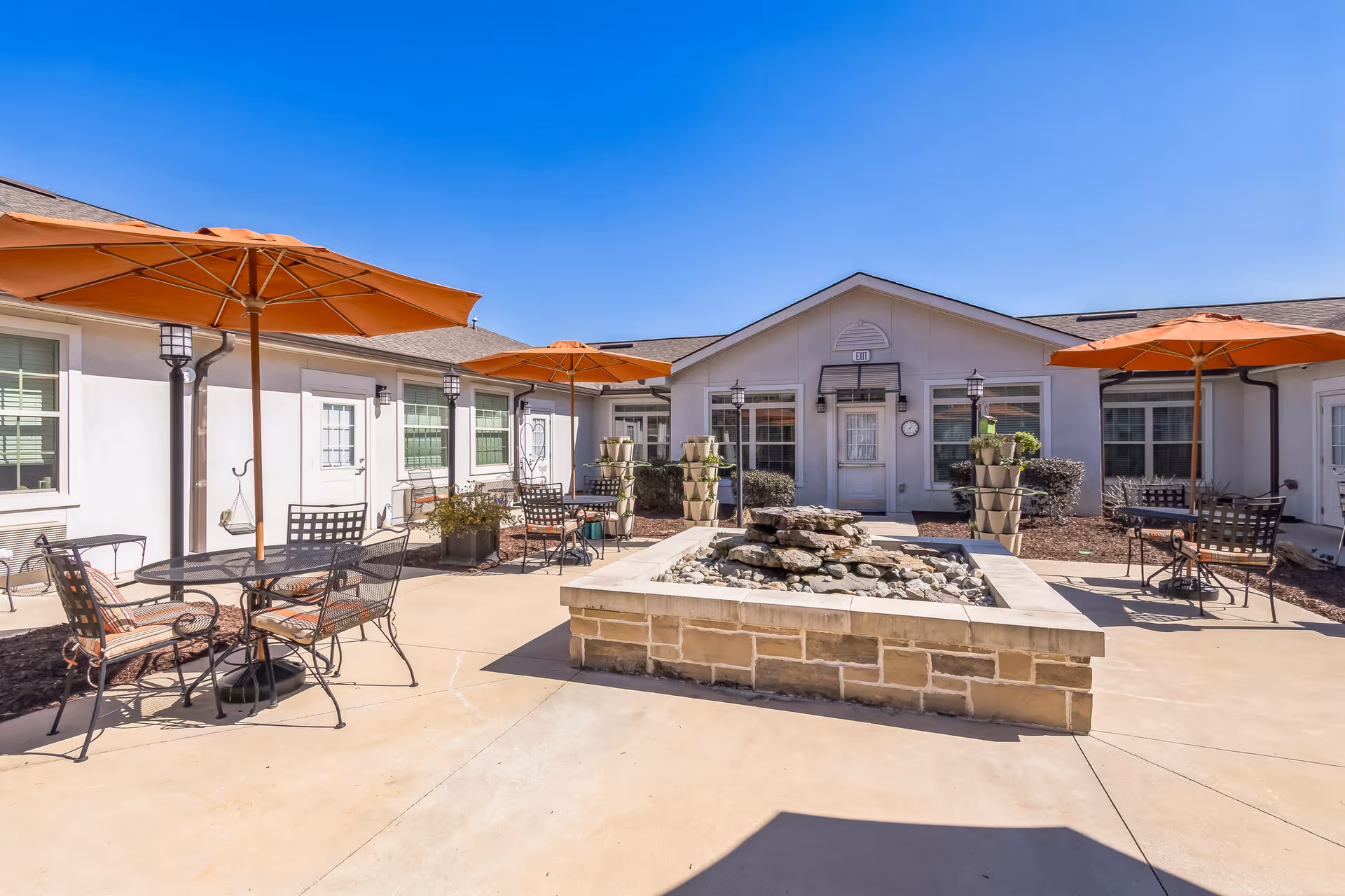 Outdoor courtyard area at Barclay House of Aiken featuring a stone water fountain in the center, surrounded by metal tables and chairs with orange umbrellas providing shade. The courtyard is enclosed by a single-story building with white walls and multiple windows and doors under a clear blue sky.