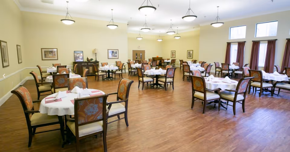 A spacious dining room with multiple round tables covered with white tablecloths and set with folded napkins. Each table is surrounded by wooden chairs with cushioned seats and backs featuring a floral pattern. The room has wooden flooring, cream-colored walls adorned with framed artwork, and several ceiling lights. Windows with dark curtains allow natural light into the room.