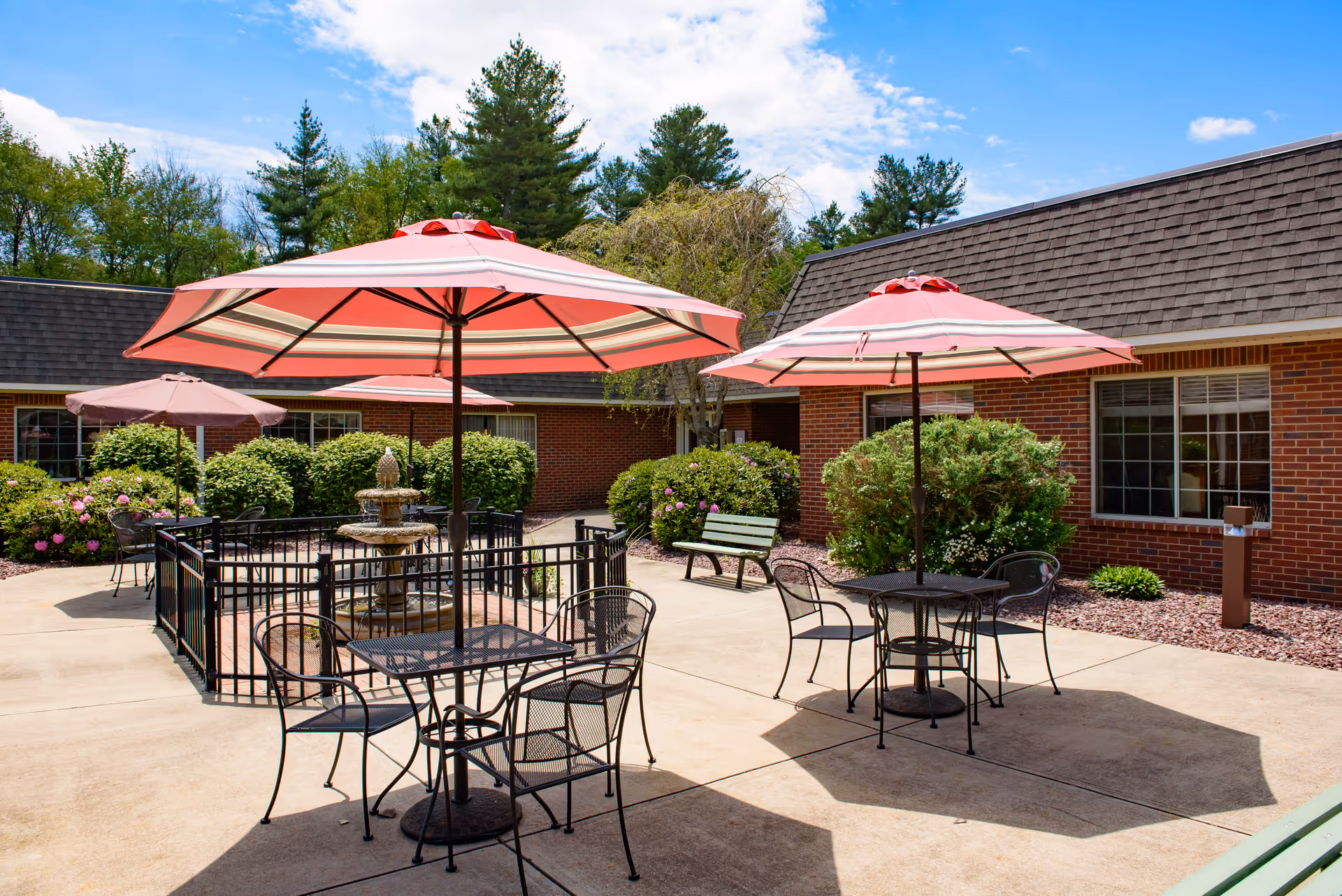 Outdoor courtyard patio with pink umbrellas, metal tables and chairs, a central fountain, and brick building in the background.