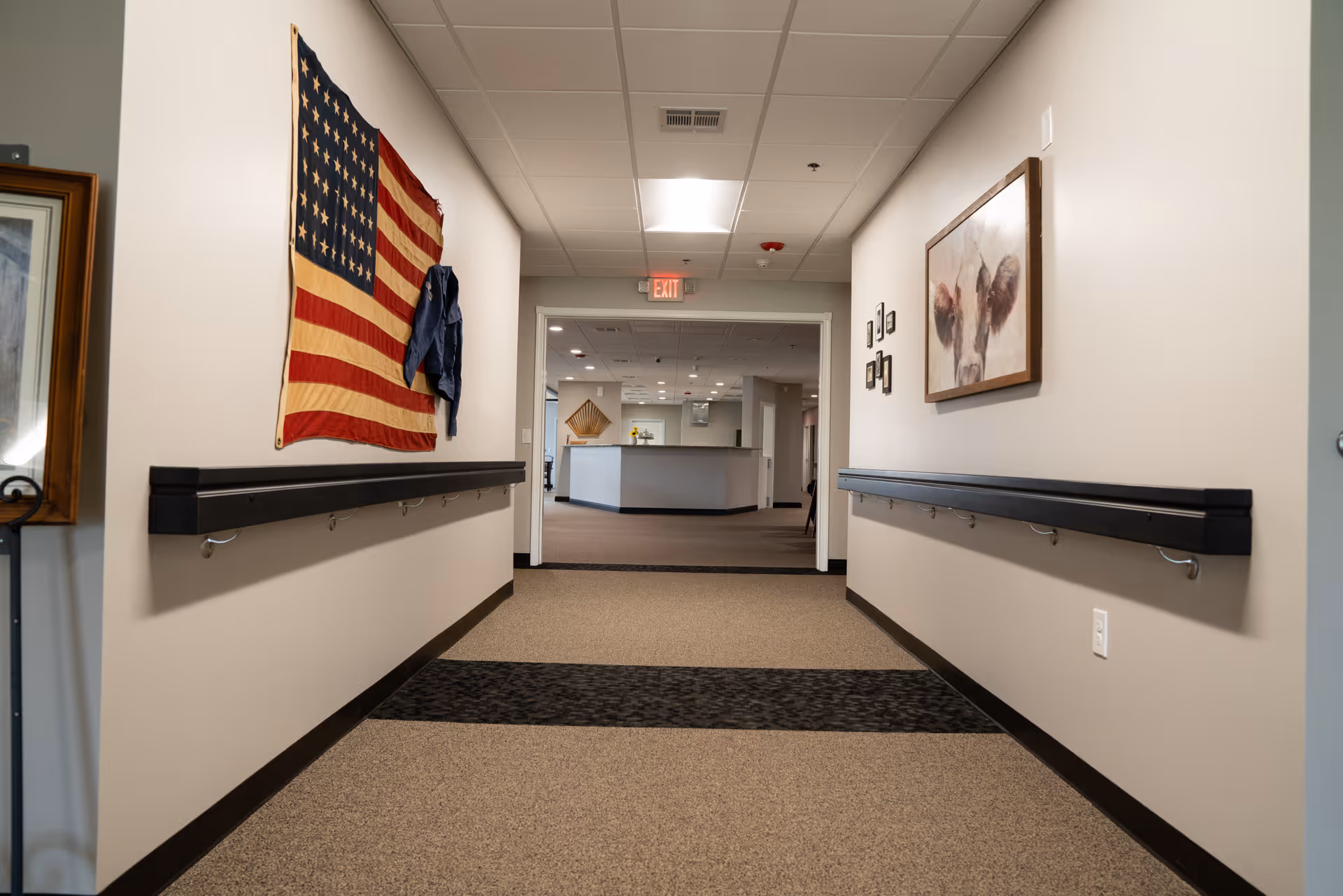 A hallway in The Mapleton Assisted Living facility with beige walls and carpeted floor. Handrails run along both sides of the hallway. An American flag and a blue jacket are hanging on the left wall, while a framed painting of a cow and several small framed pictures are on the right wall. The hallway leads to a reception or common area with a counter and ceiling lights.