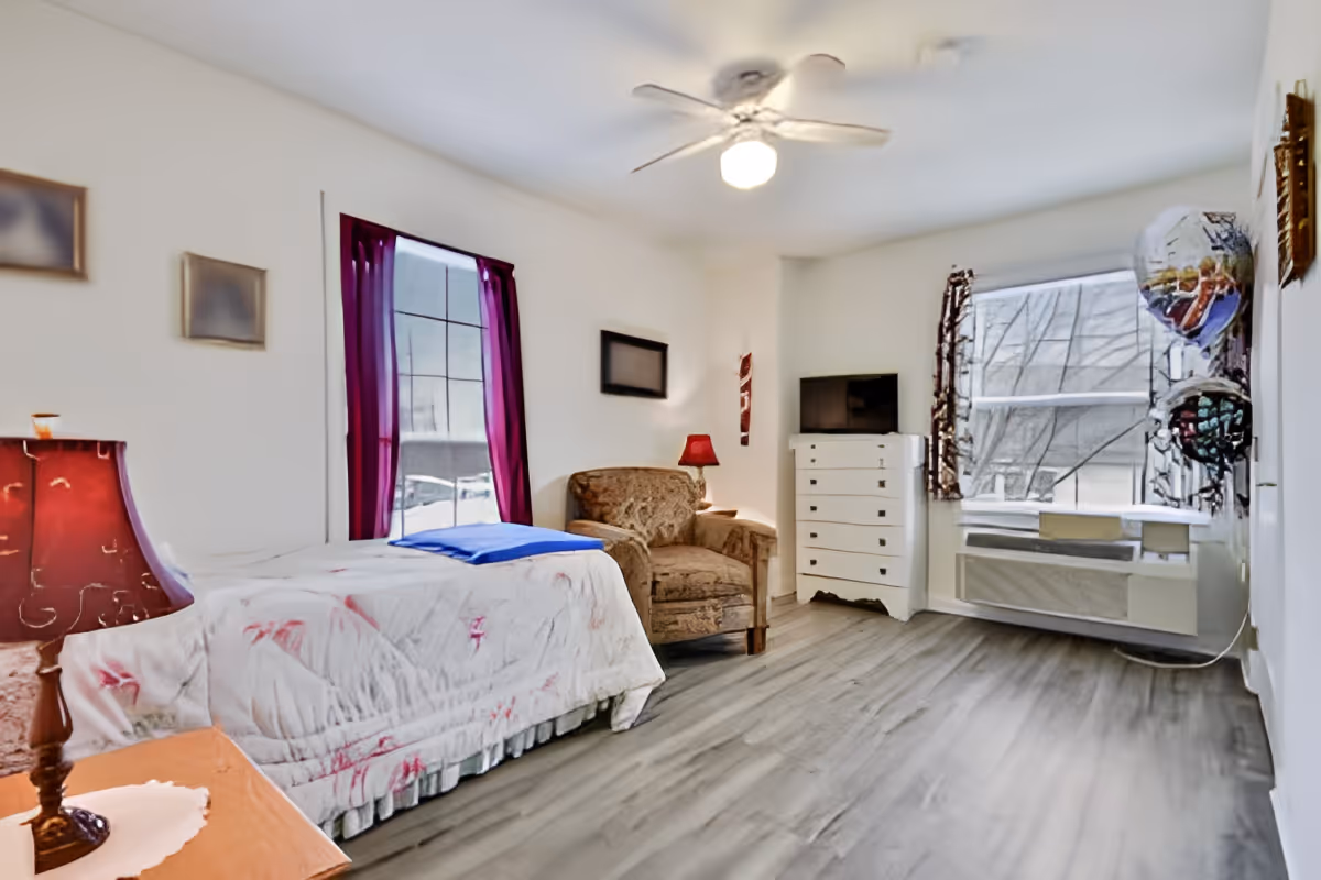 Bright single-occupancy bedroom with a bed, armchair, dresser and TV beneath windows and a ceiling fan.