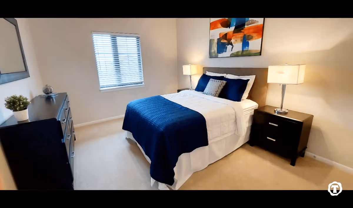 A neatly arranged bedroom with a bed covered in white and blue bedding, two bedside tables each with a lamp, a dresser with a small plant and decorative item, a window with blinds, and a colorful abstract painting on the wall above the bed.