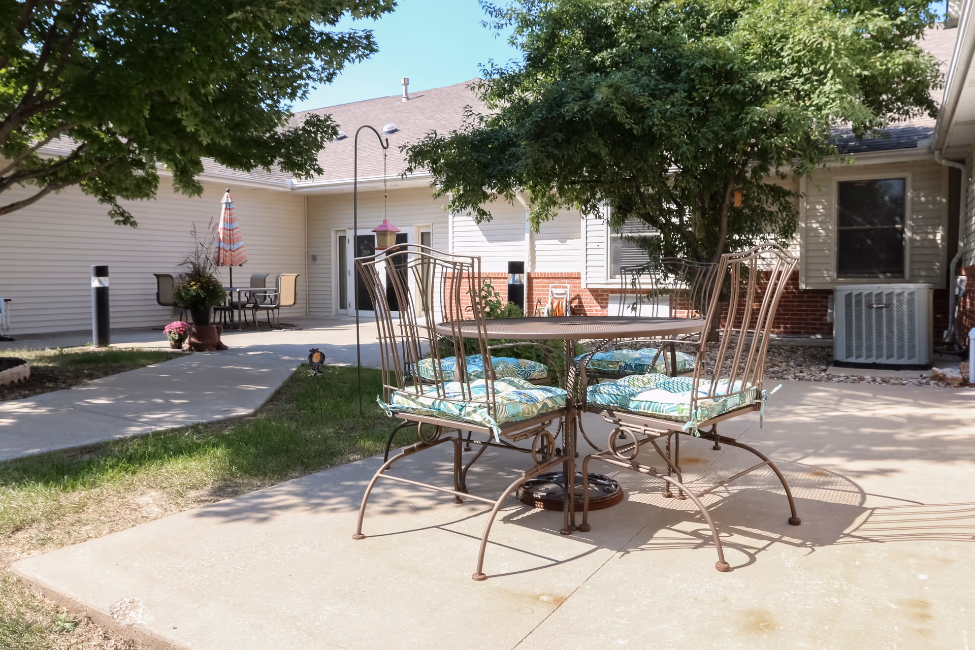 Outdoor patio area with a metal table and four chairs with patterned cushions, surrounded by trees and adjacent to a building with white siding and brick accents. There is another seating area with a table and umbrella in the background.