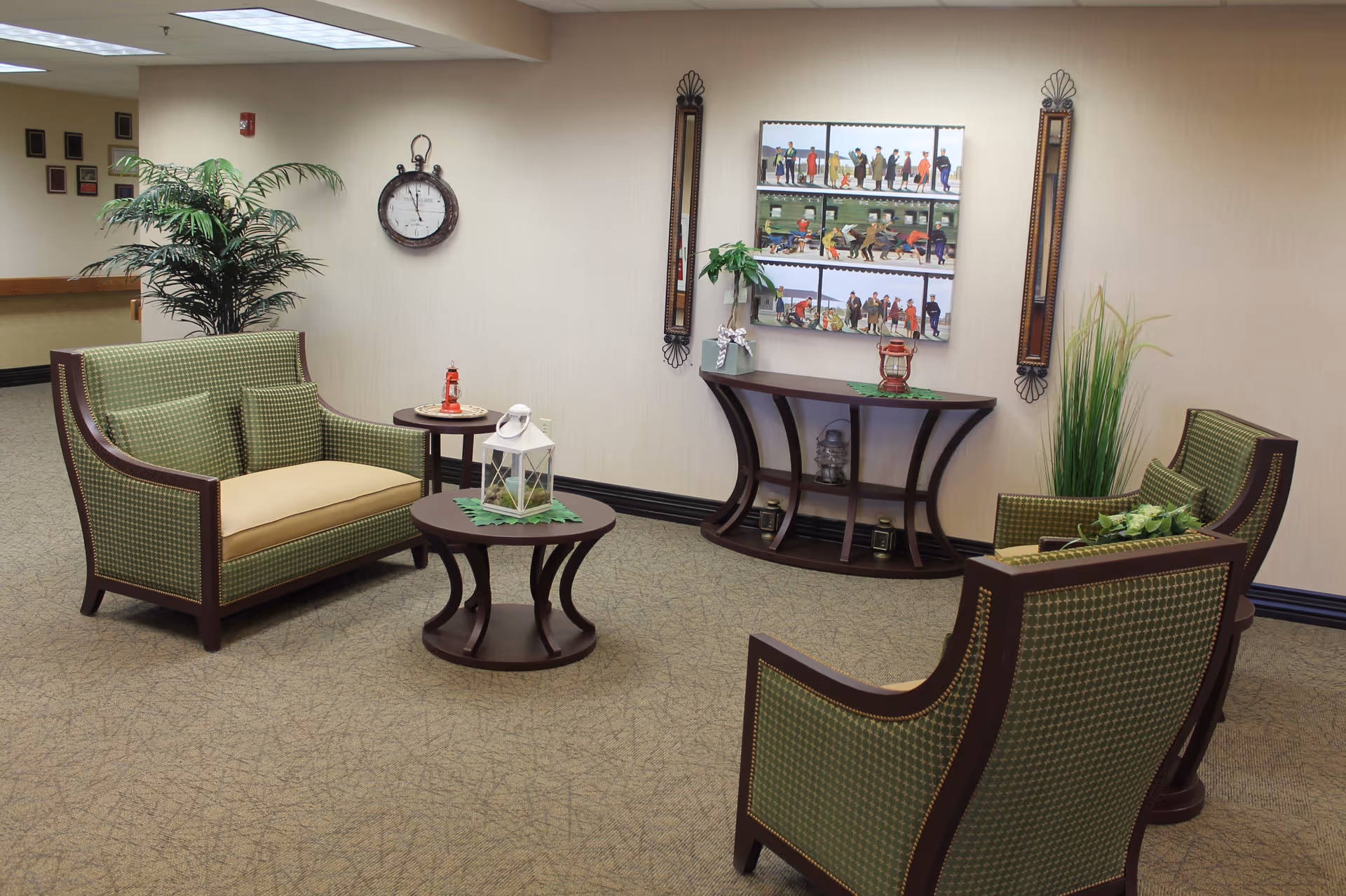 A cozy sitting area in a senior living facility with green cushioned chairs and a loveseat arranged around two small wooden tables. The tables hold decorative lanterns and plants. The walls are beige with framed artwork and a clock, and there are tall plants in the corners.