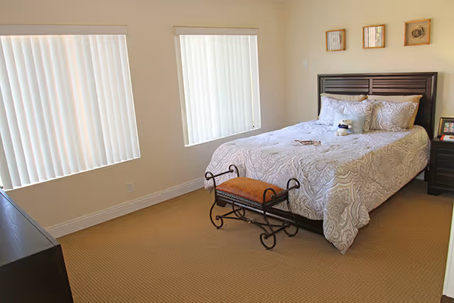 A bright bedroom featuring a double bed with patterned bedding, a decorative bench at the foot, a nightstand, and two windows with vertical blinds.