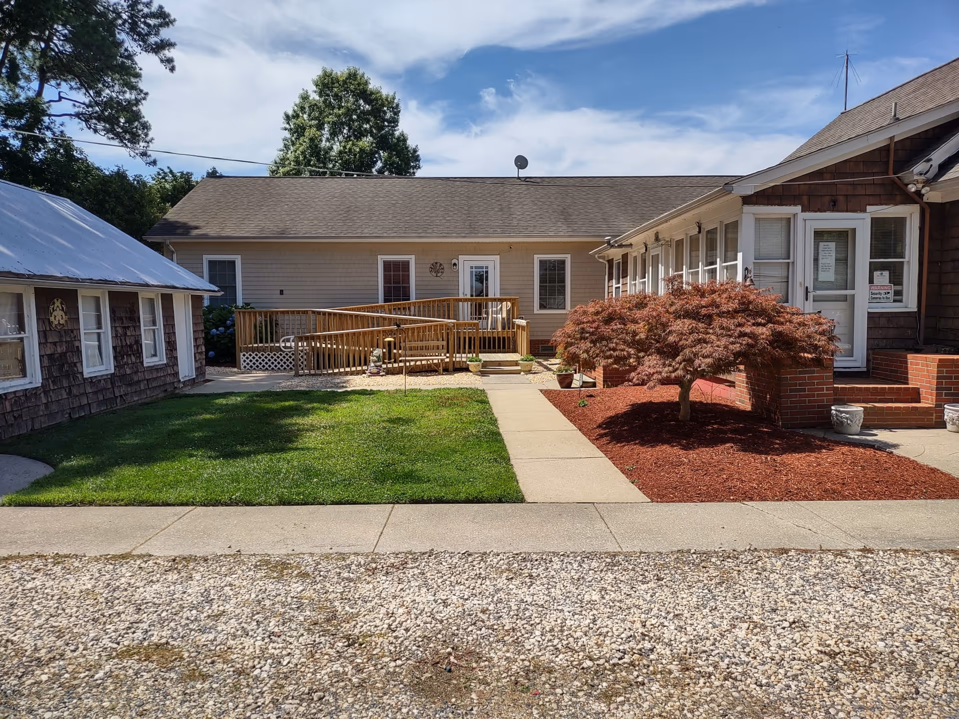 Exterior view of Whispering Pines Assisted Living Facility showing a single-story building with a ramp and stairs leading to the entrance. There is a well-maintained lawn, a small tree with red leaves, and a gravel area in the foreground under a partly cloudy sky.
