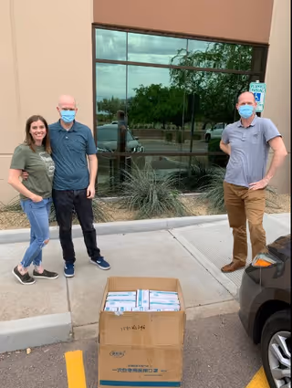Three people wearing face masks standing outside a building near a large cardboard box filled with supplies. The building has a large window reflecting trees and cars in the parking lot.