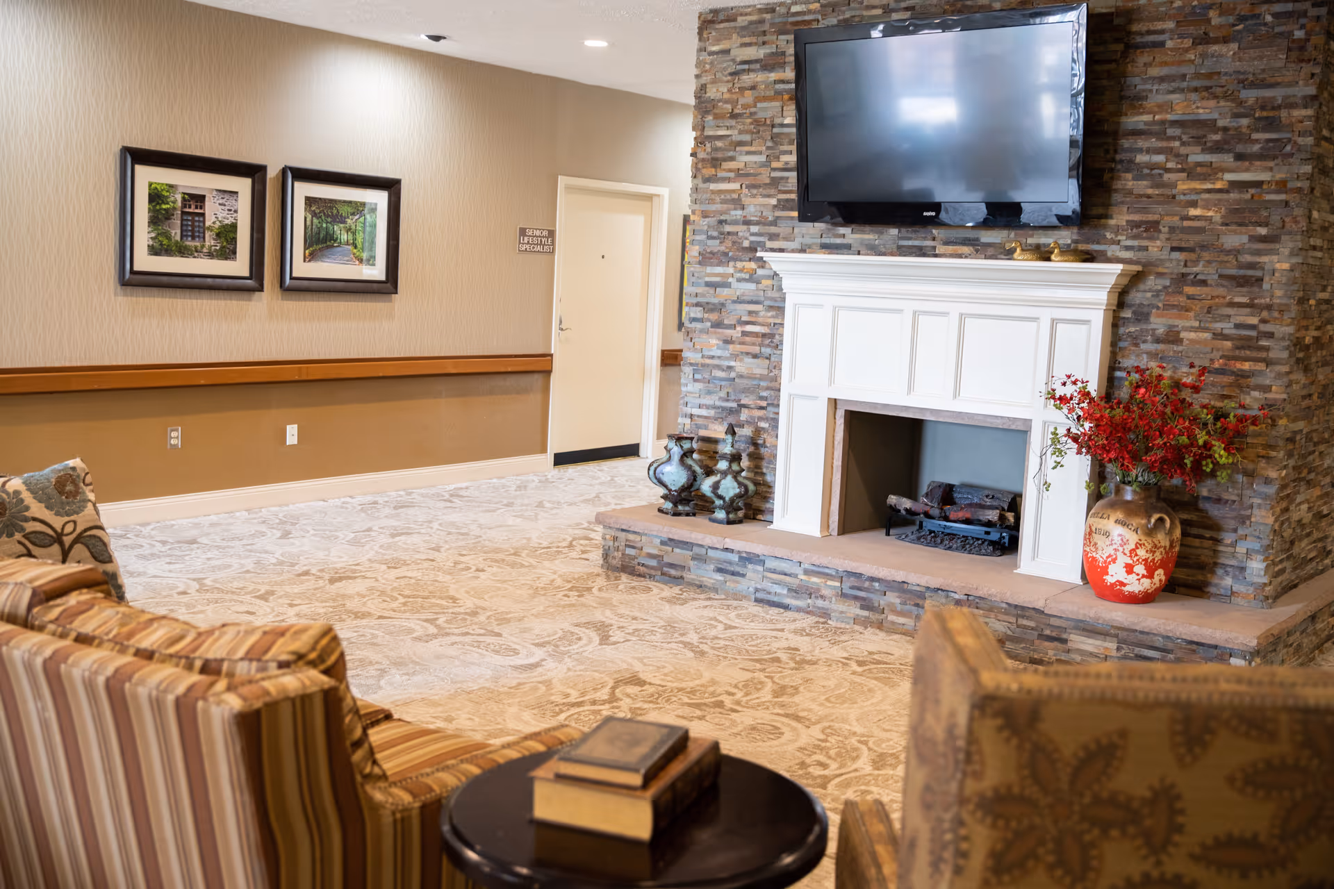 A cozy living room area featuring a stone fireplace with a white mantel, a flat-screen TV mounted above it, decorative vases on the hearth, and a large vase with red flowers. The room has patterned carpet, striped and floral upholstered chairs, a small round table with books, and framed pictures on the beige wall.