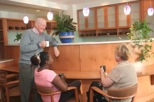 Three people socializing in a kitchen area; an elderly man standing and holding a white mug, and two women sitting on chairs holding black mugs, with wooden cabinets, hanging pendant lights, and green plants in the background.