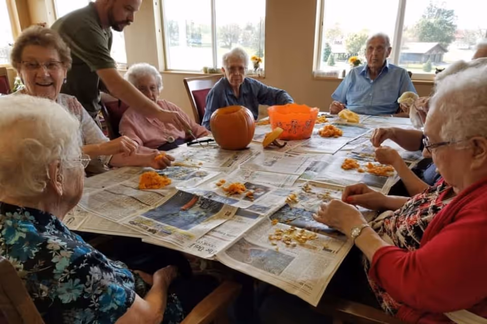 A group of elderly people sitting around a table covered with newspapers, engaged in scooping out pumpkin seeds and pulp. A man is assisting them. There is a carved pumpkin and an orange plastic container on the table. The room has large windows letting in natural light.