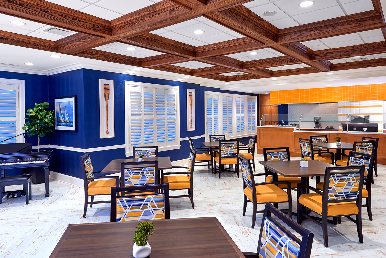A bright dining room with multiple square wooden tables and chairs featuring yellow cushions and geometric patterned backs. The room has blue walls with white trim, decorative wooden ceiling beams, and white plantation shutters on the windows. A black grand piano is positioned in the corner next to a green plant and framed nautical artwork. The back of the room has a serving counter with a yellow tiled backsplash.