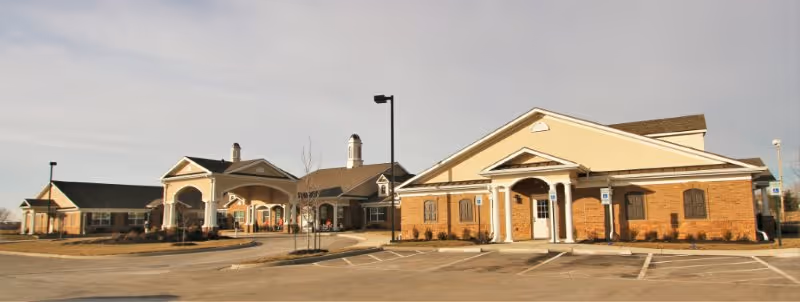Wide exterior view of a single-story brick and stucco senior living facility with arched entrances and an empty parking lot.