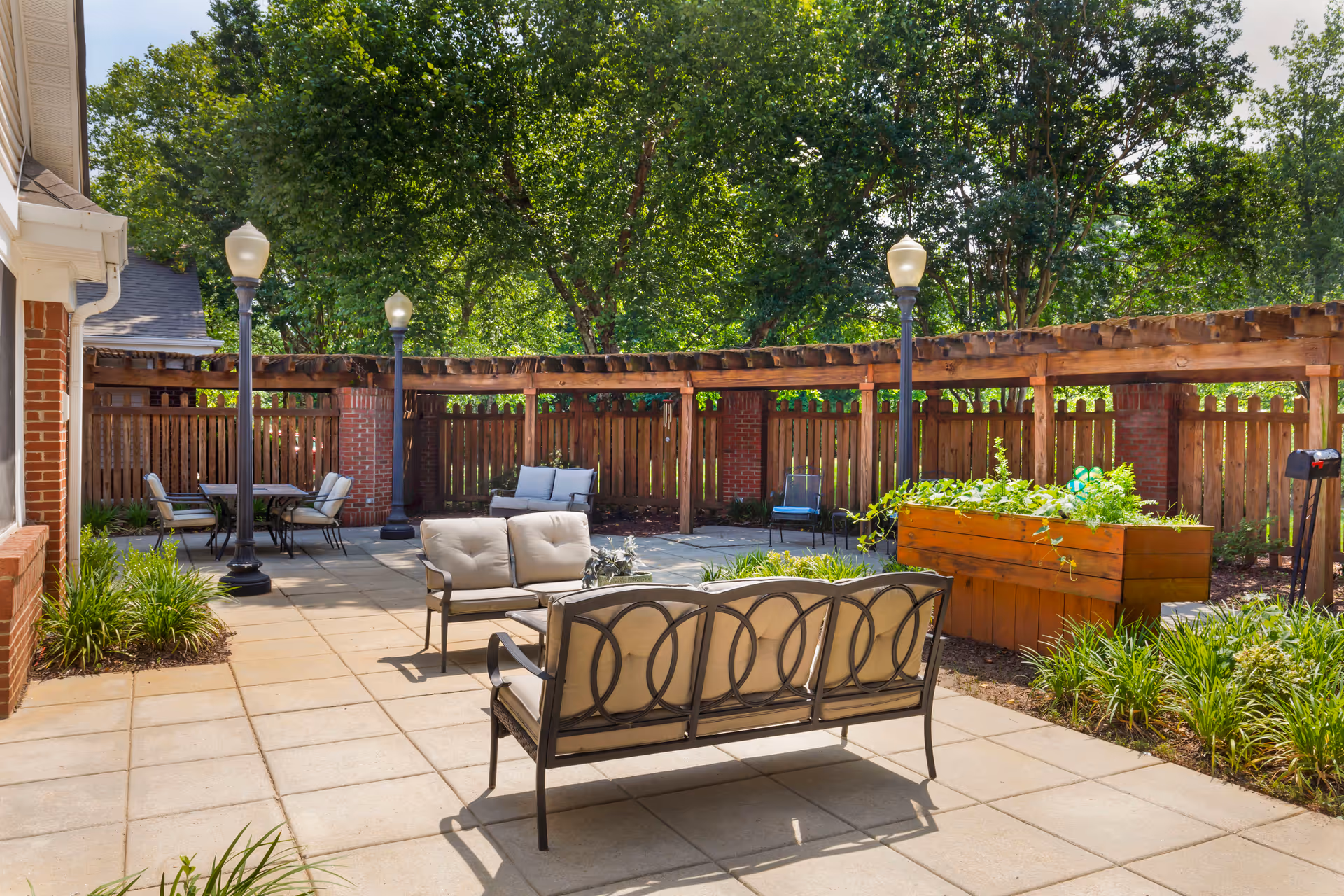 Outdoor patio area with cushioned metal seating, a wooden planter box with greenery, several lamp posts, and a wooden fence with brick pillars surrounded by trees.