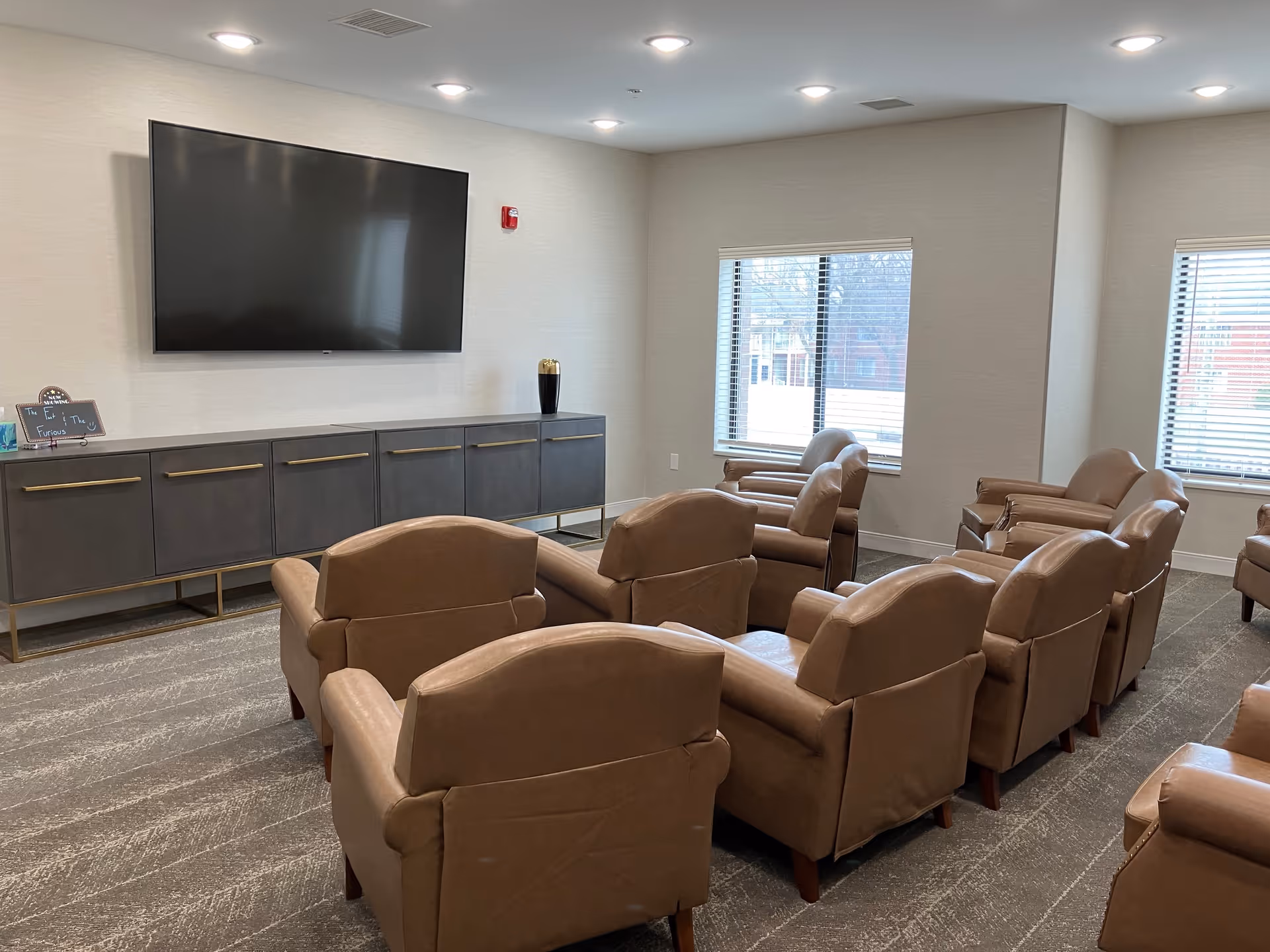 A common area with multiple brown leather armchairs arranged in rows facing a large flat-screen TV mounted on a light-colored wall. Below the TV is a long dark gray cabinet with gold handles. Two windows with blinds allow natural light into the room.