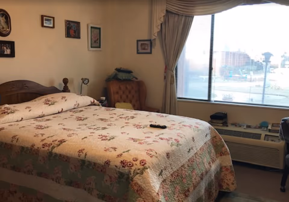 A bedroom with a bed covered in a floral quilt, a wooden headboard, and a remote control on the bed. There is a cushioned armchair with pillows in the corner near a large window with blinds and curtains. Several framed pictures hang on the wall above the bed.