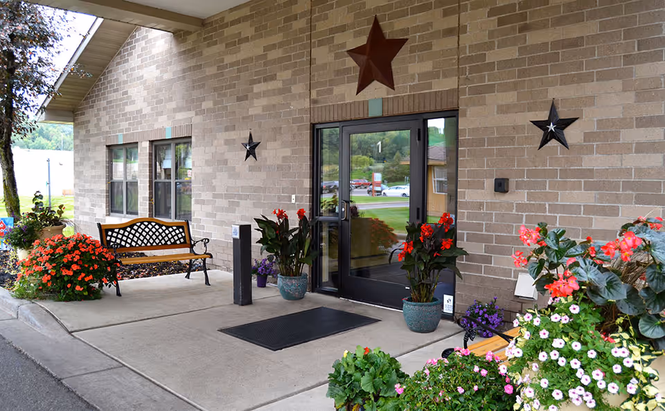 Covered front entrance of a brick nursing home with glass double doors, benches, and potted flowering plants.