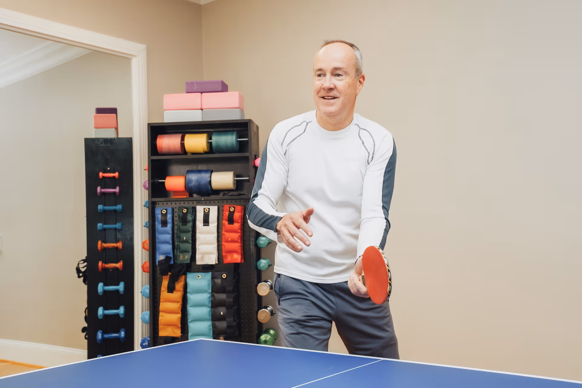 A man in a white long-sleeve shirt and gray pants playing table tennis indoors. Behind him is a rack holding various exercise equipment including dumbbells, resistance bands, and yoga blocks. The room has beige walls and a large mirror on the left side.