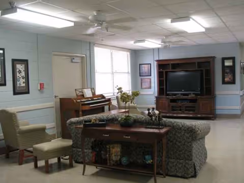 A communal living room in a senior facility with sofa, piano, TV, and plants.