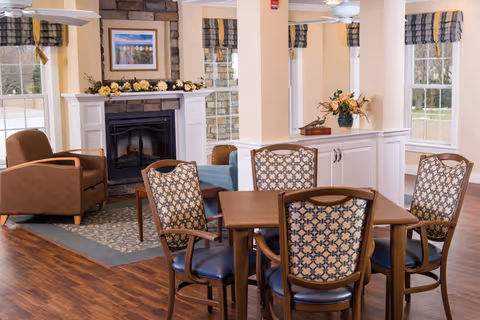 A cozy interior common area with a wooden table and four patterned chairs in the foreground. Behind the table is a white cabinet with a flower arrangement on top. To the left, there is a stone fireplace with a framed picture above it and a brown armchair nearby. The room has large windows with plaid valances, light-colored walls, and wood flooring.