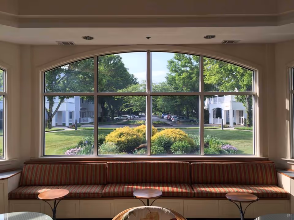 Sunlit lounge with striped bench seating and small tables facing a large arched window overlooking landscaped grounds and parked cars.