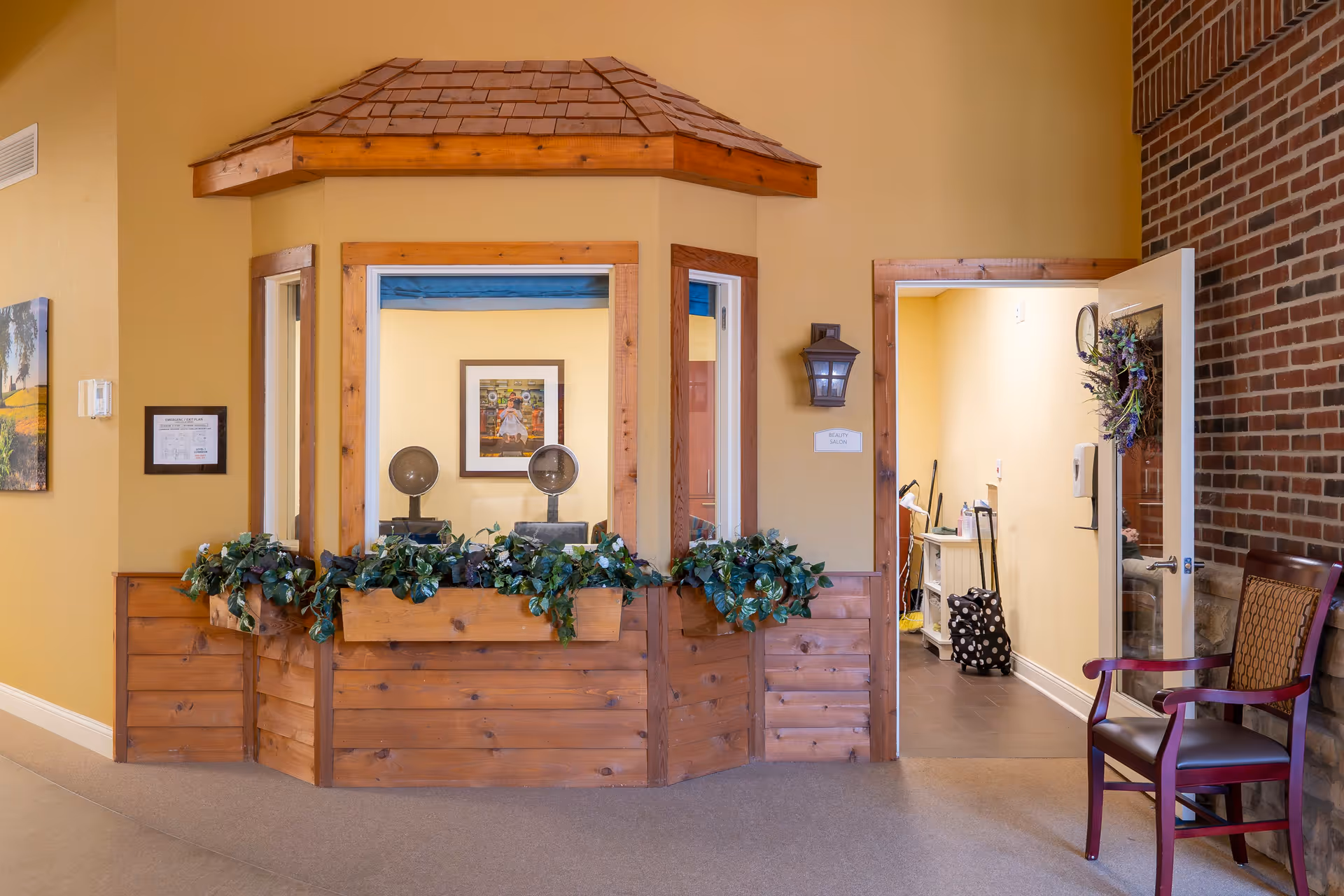Interior view of a reception or check-in area with a wooden counter and a small roof structure above it. The counter has green leafy plants along the front. Behind the counter are two hair dryers and a framed picture on the wall. To the right, there is an open door leading to a small room with cleaning supplies and a chair with a patterned backrest next to a brick wall.