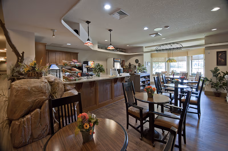 Bright dining room with wooden tables and chairs, a serving counter, and decorative plants and lighting.