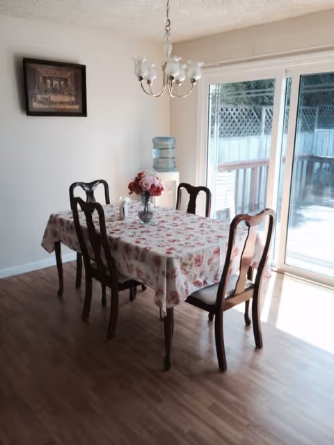 A dining area with a rectangular wooden table covered with a floral tablecloth and surrounded by four wooden chairs. A vase with flowers is placed in the center of the table. There is a water dispenser against the wall and a framed picture hanging above it. Sliding glass doors lead to an outdoor deck with a white fence visible outside.