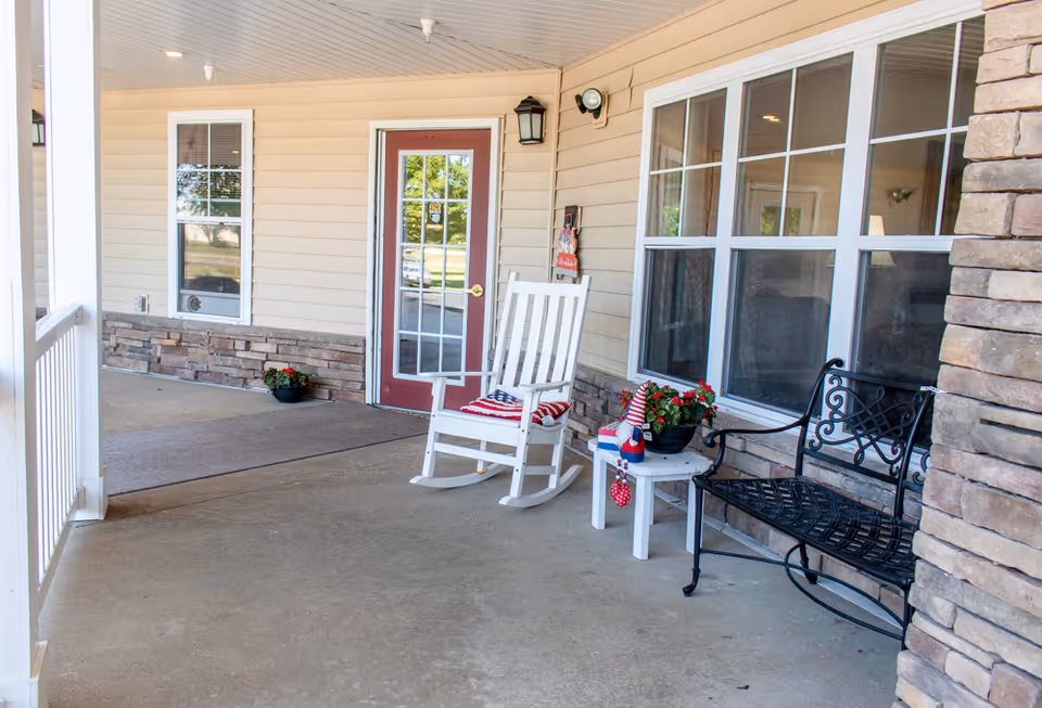 Covered front porch with a white rocking chair, metal bench, potted flowers and a glass-paned entry door.