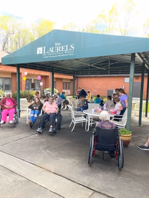 Residents and staff sit and socialize under a green entrance canopy labeled 'The Laurels of GreenTree Ridge' outside a brick senior living facility.