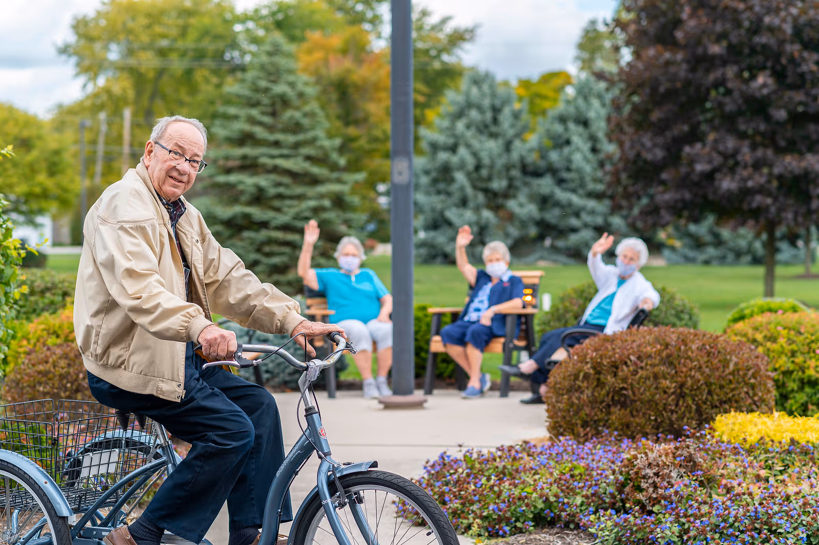 An elderly man wearing glasses and a beige jacket is sitting on a bicycle outdoors in a garden area. In the background, three elderly women wearing face masks are sitting on benches and waving. The scene is set in a green, landscaped area with trees, bushes, and flowers.